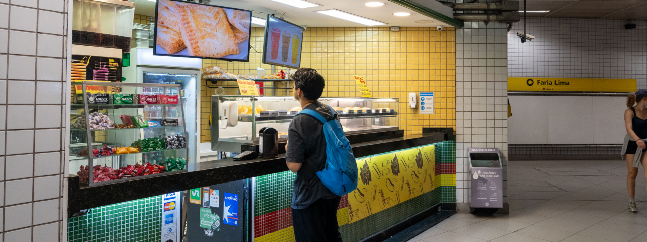 Jovem com mochila azul em pé diante de uma lanchonete na Estação Faria Lima do Metrô de São Paulo.
