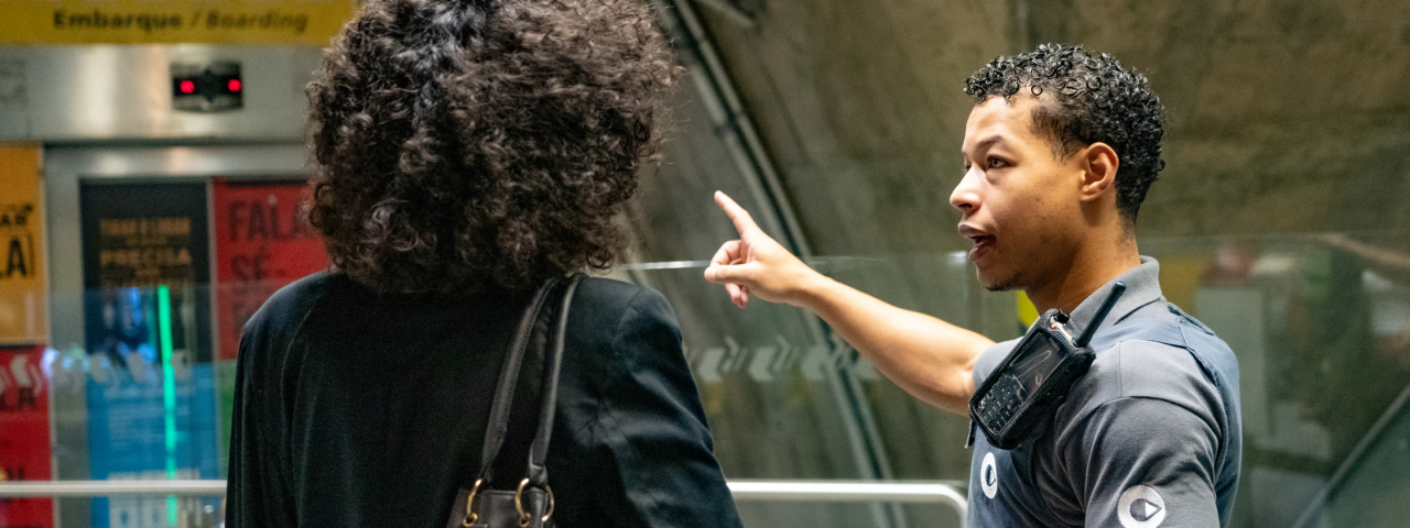 Agente de estação orientando passageira, apontando direção em área de embarque com placas informativas ao fundo.