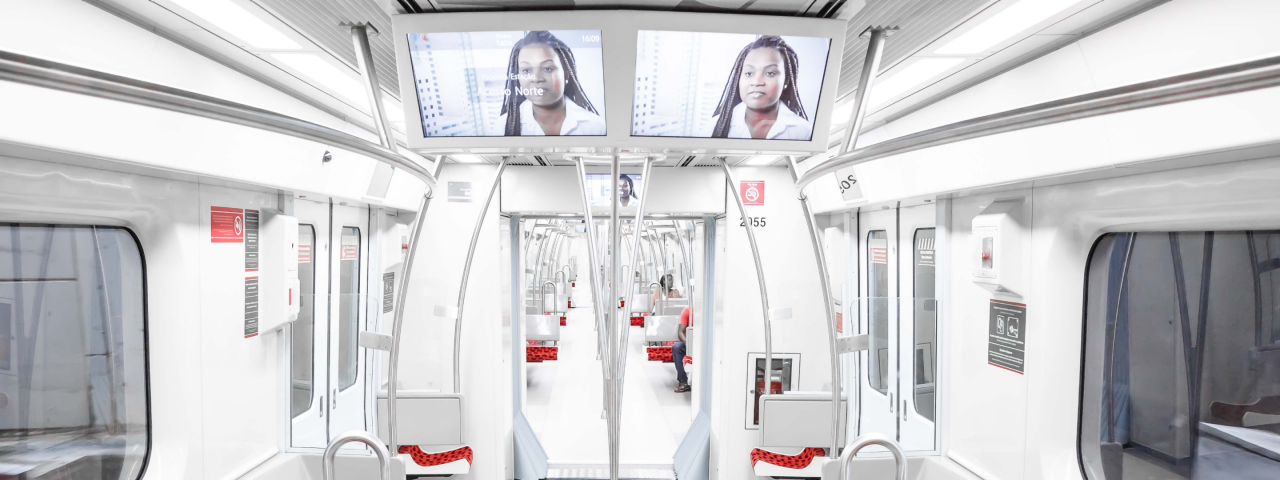 Imagem mostrando o interior de um vagão de metrô na cor branca com detalhes vermelhos com três passageiros ao fundo.