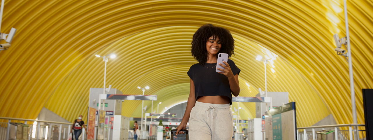 Mulher com celular nas mãos, sorrindo diante da entrada da estação.