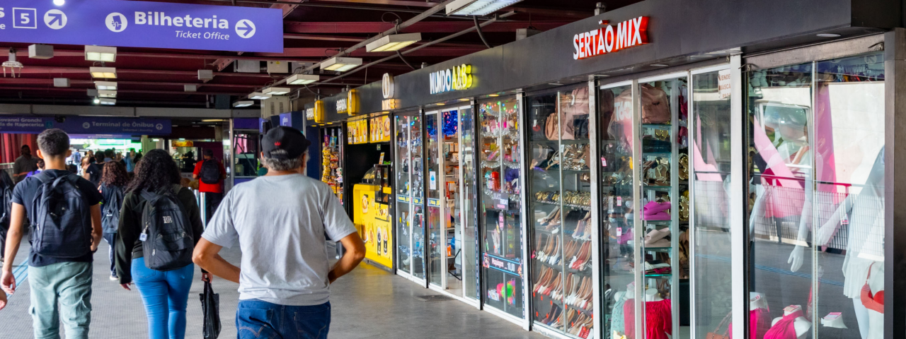 Passageiros caminhando em estação com lojas e bilheteria, fachada com letreiro Sertão Mix e Mundo ABB visíveis.
