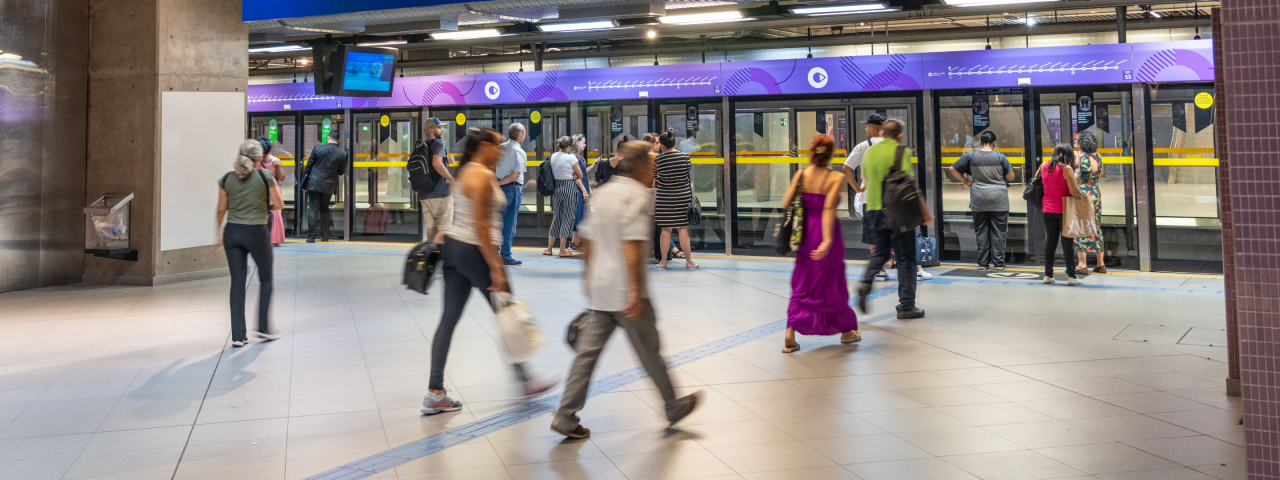 Passageiros aguardam na plataforma do metrô em frente às portas de vidro de proteção.