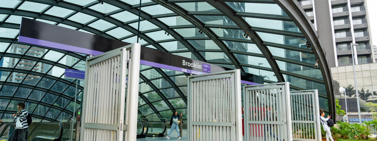 Entrada da estação de metrô Brooklin com cobertura de vidro e pessoas circulando na área externa.