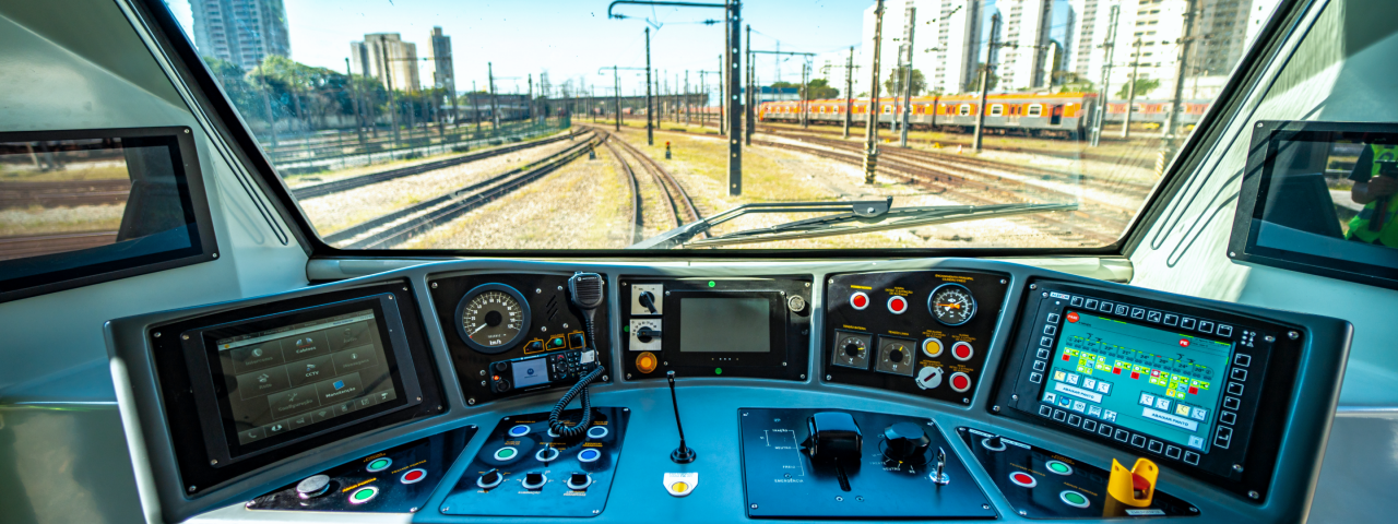 Cabine de comando de um trem, oferecendo uma visão detalhada do painel de controle e da paisagem ferroviária à frente.