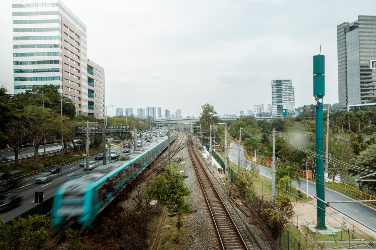 Vista aérea de trem da Linha 9-Esmeralda percorrendo os trilhos paralelos à Marginal Pinheiros e edifícios corporativos.