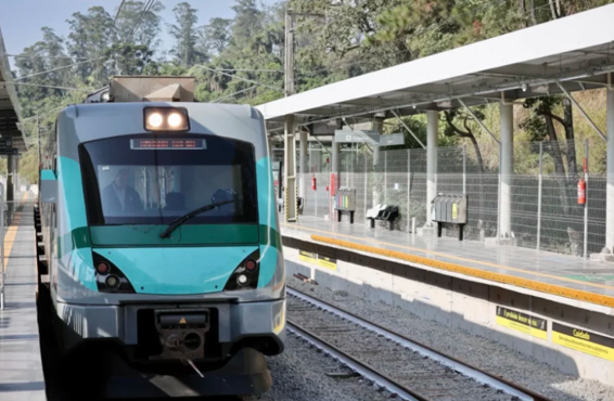 Trem turquesa e cinza se aproxima de plataforma coberta cercada por vegetação e mata