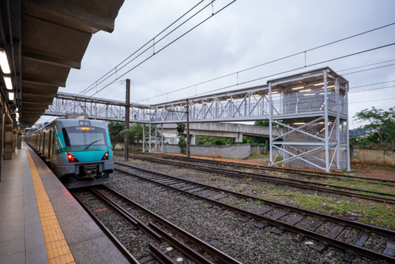 Trem parado em plataforma com passarela metálica elevada e torre de elevador ao fundo, em dia nublado