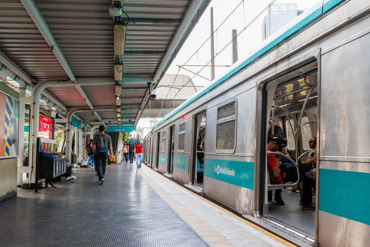Passageiros caminhando pela plataforma coberta de uma estação da Linha 9-Esmeralda da ViaMobilidade ao lado do trem com portas abertas.