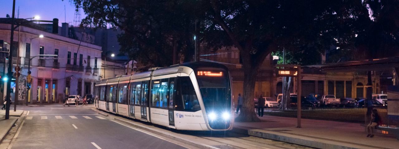 VLT do Rio de Janeiro à noite na estação, iluminado pelos faróis, com prédios históricos e árvores ao redor.